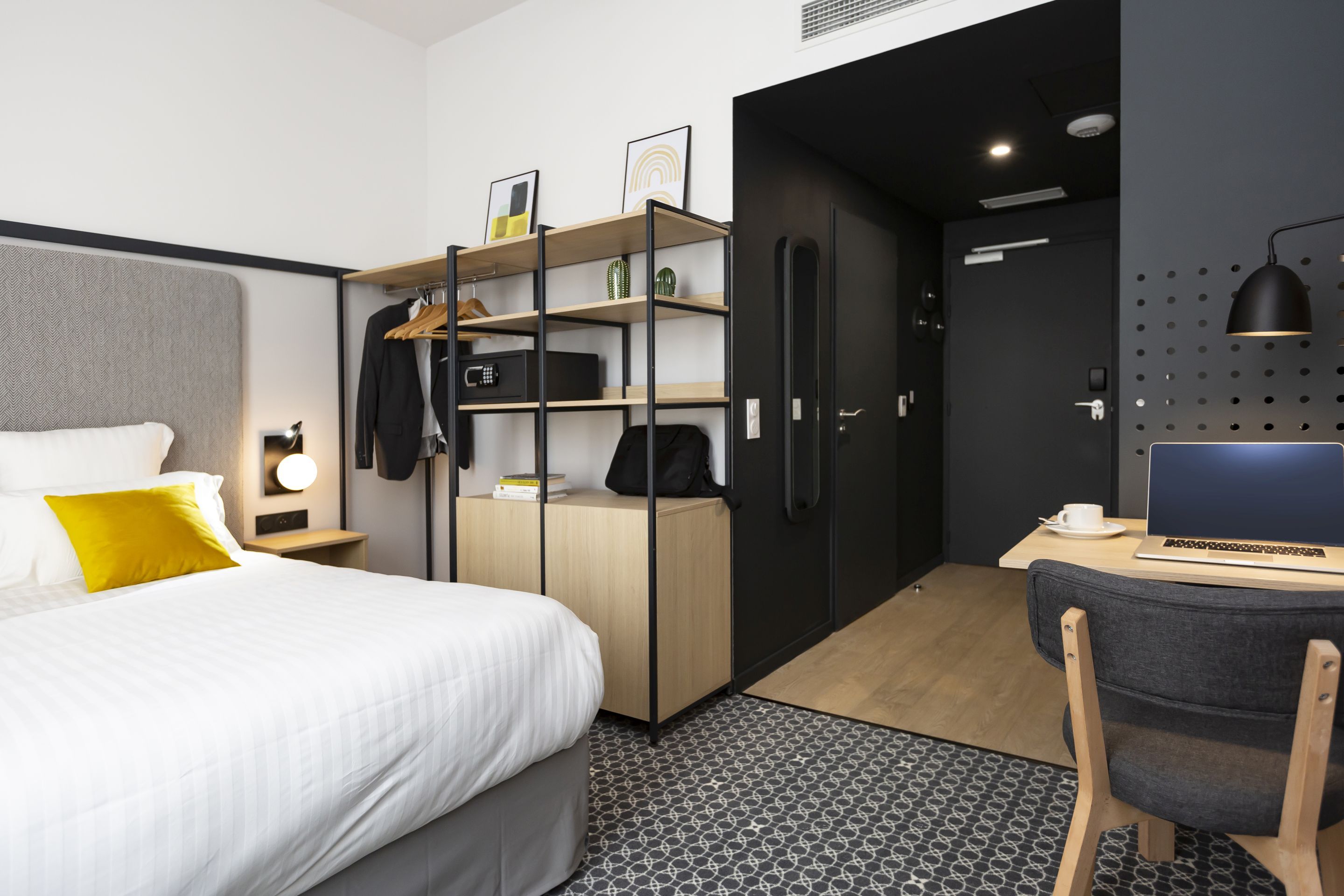 A modern hotel room with a bed, a desk with a laptop, a wardrobe, and a shower area.