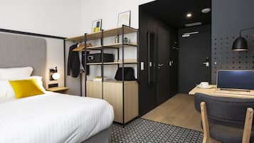 A modern hotel room with a bed, a desk with a laptop, a wardrobe, and a shower area.