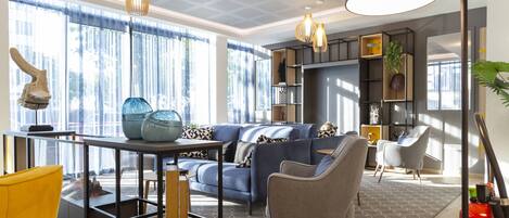 Una sala de estar moderna con un sofá azul, un sillón amarillo y una mesa de centro de vidrio. Hay lámparas colgantes, una estantería y un ventanal con cortinas.