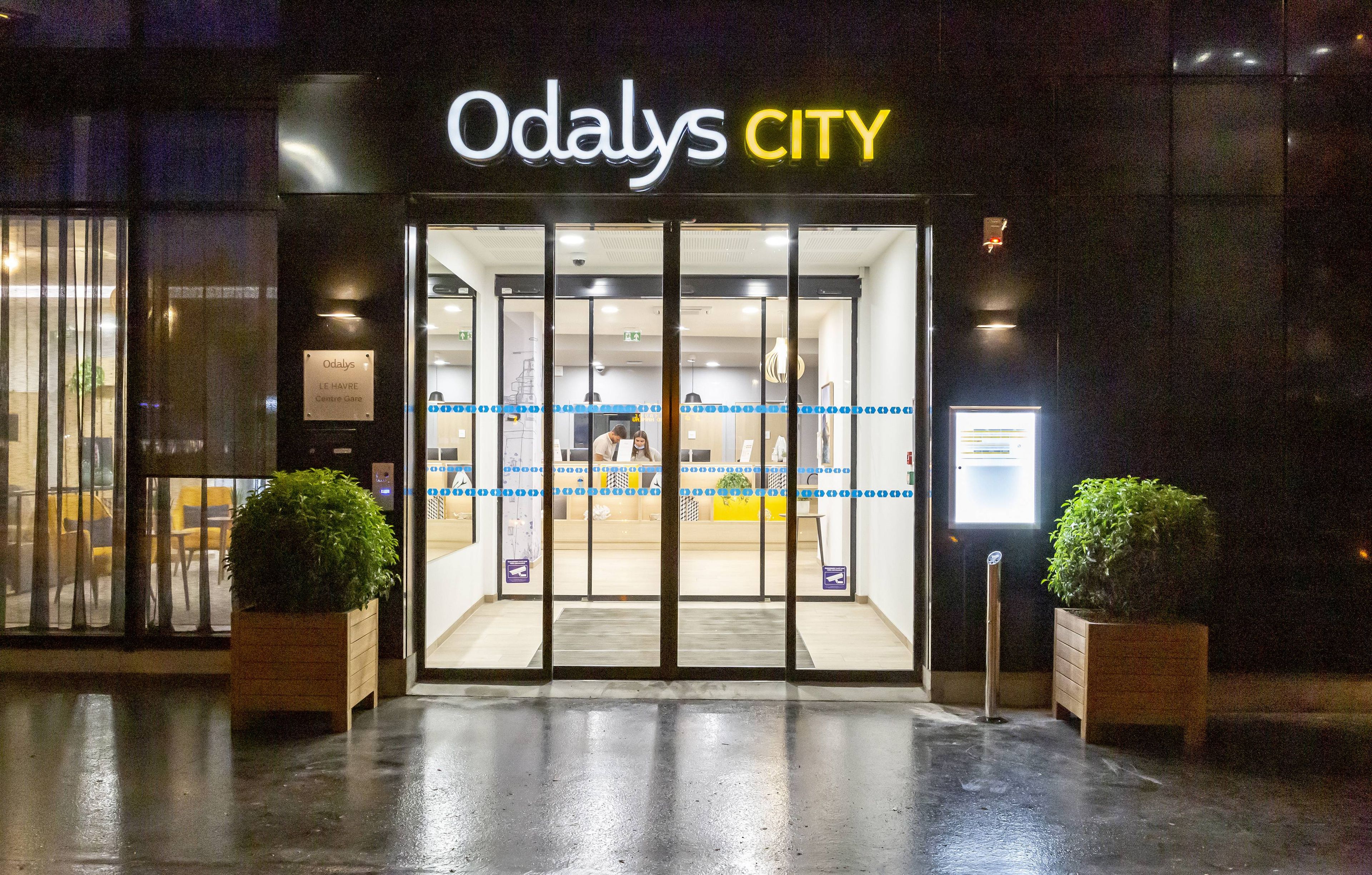 The entrance to Odalys CITY with a glass door, a sign above the door, and two potted plants on either side.