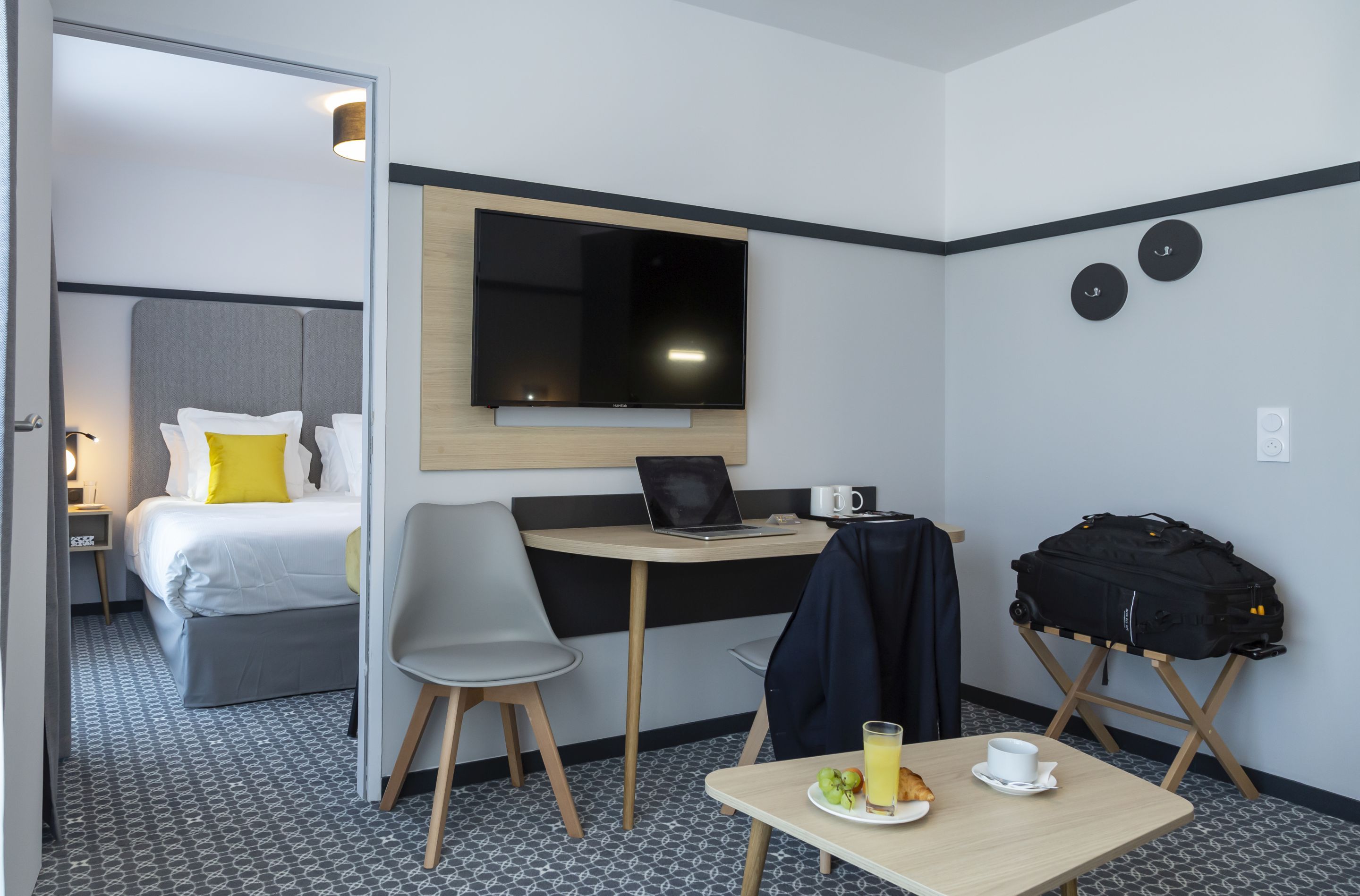 Habitación de hotel con cama, escritorio con una computadora portátil, silla, mesa con desayuno y maleta.