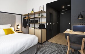 Une chambre d’hôtel moderne équipée d’un lit, d’un bureau avec un ordinateur portable, d’une armoire et d’un petit coin salon.