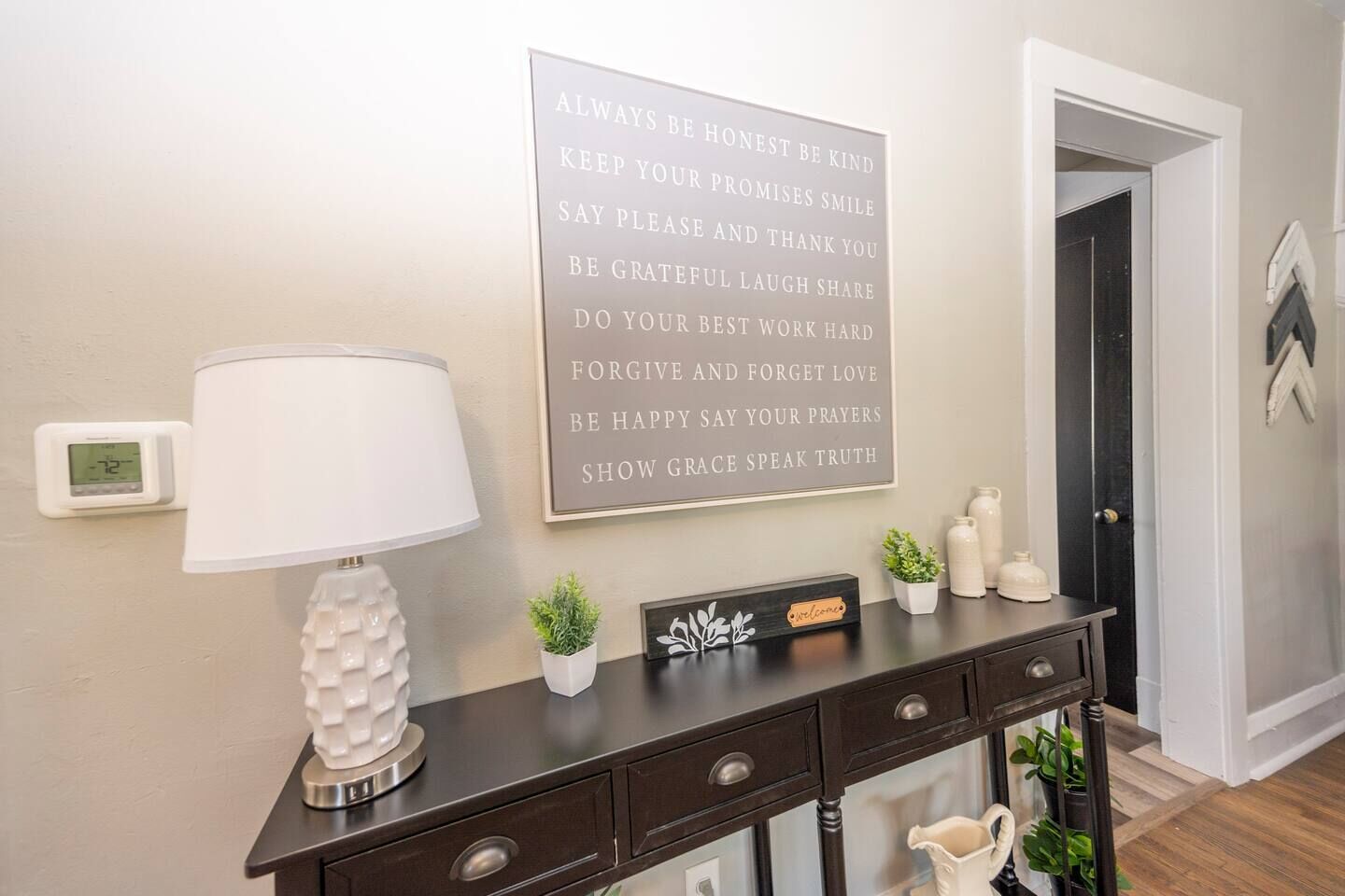 A wall with a motivational quote, a digital thermostat, a lamp, a small plant, and a welcome sign on a dark wooden console table.