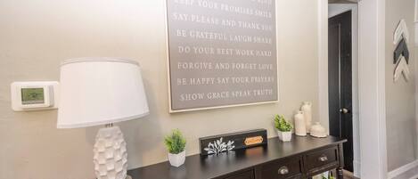 A wall with a motivational quote, a digital thermostat, a lamp, a small plant, and a welcome sign on a dark wooden console table.