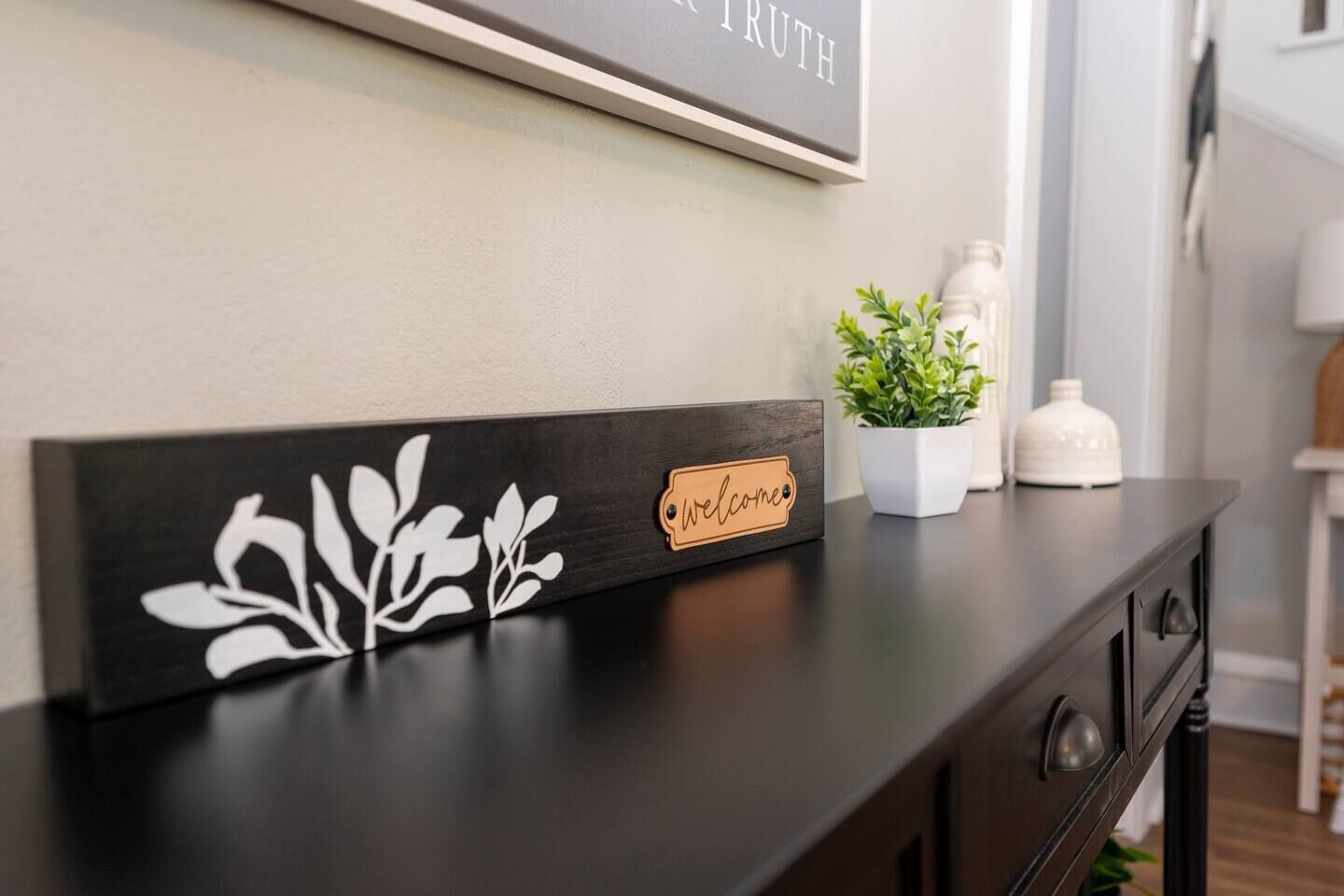 Une console en bois foncé, ornée d’une plaque décorative sur laquelle est inscrit le mot « bienvenue », et au-dessus, un cadre contenant l’inscription « EN VÉRITÉ ».