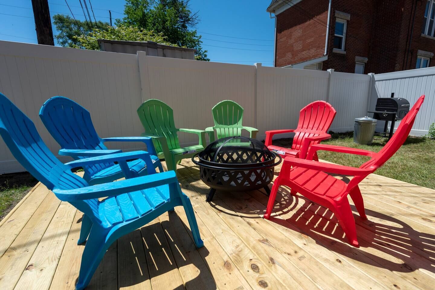 Un patio trasero con una terraza de madera, un brasero y sillas Adirondack de colores.