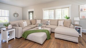 Un salon avec un canapé d’angle, une petite table d’appoint, un plaid vert et une horloge au mur.