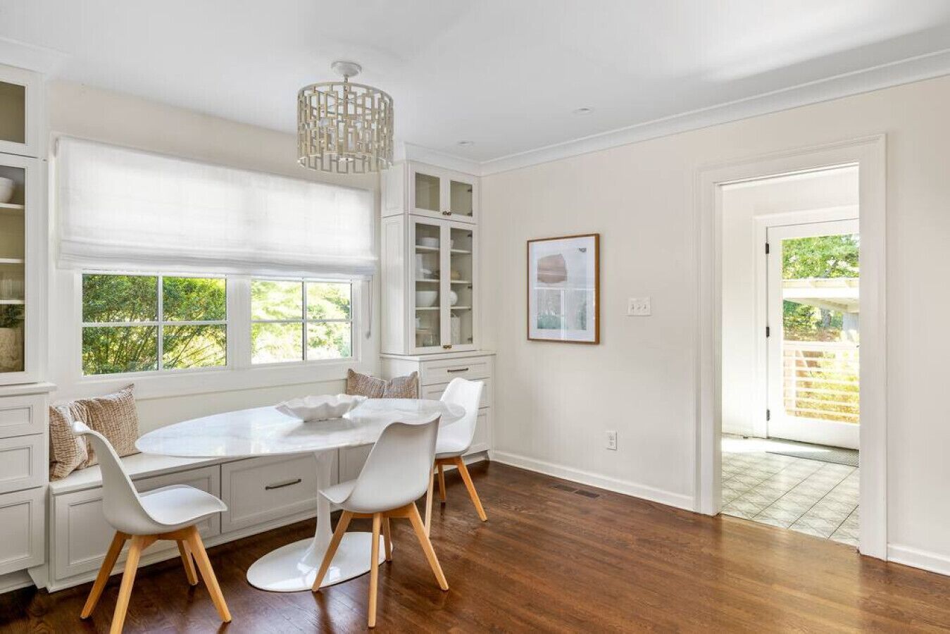 Un espace repas avec une table et des chaises blanches, un lustre et une vue sur la verdure à l’extérieur.