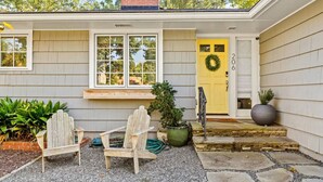 Ein Haus mit gelber Haustür, zwei Fenstern, einer kleinen Veranda mit zwei Adirondack-Stühlen und einem Blumentopf.