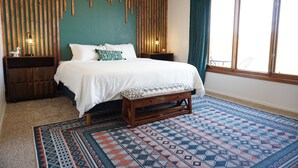 A bedroom with a bed, wooden headboard, bedside tables, a bench, a patterned rug, and a large window with curtains. - Unleash Your Inner Explorer: The Dam Dino House! (Page)