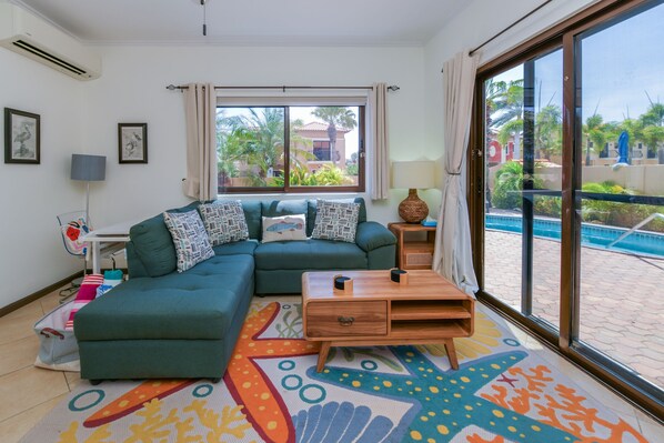 A living room with a sectional sofa, a wooden coffee table, a colorful rug, a window with curtains, and a view of a swimming pool.