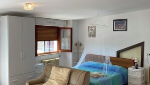 Een slaapkamer met een bed, een stoel, een nachtkastje, een radiator, een raam met zonwering, een wandplank en een plafondlamp.
