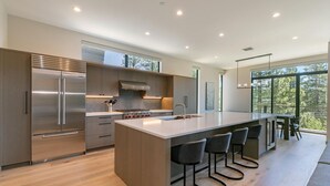 A modern kitchen with a large island, stainless steel appliances, and a view of trees through large windows. - Larkspur Luxe - Northstar- 4 BR w Pool, Gym, Access - Walk to Village. Hot Tub! (Truckee)
