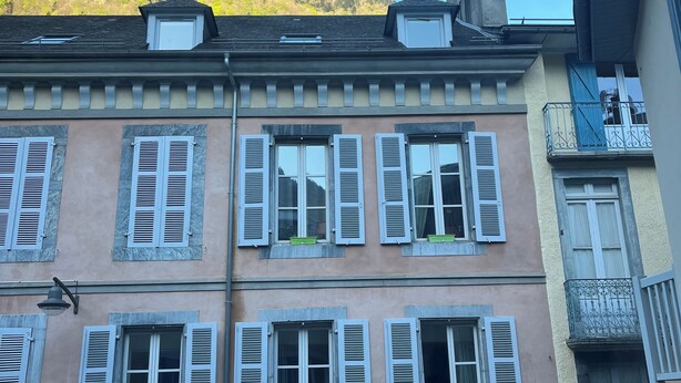 A multi-story building with pastel-colored facades, multiple windows with shutters, and a balcony with a wrought iron railing.