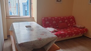 A room with a table covered in a floral tablecloth, a red sofa with patterned cushions, and a window with curtains.
