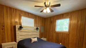 Un dormitorio con paneles de madera, una cama con colcha azul oscuro, un ventilador de techo y dos ventanas con persianas.