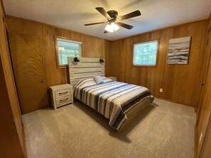 Une chambre à coucher comprenant un lit, des tables de chevet, un ventilateur de plafond et deux fenêtres équipées de stores.