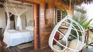 A wooden bedroom with a bed under a white canopy, a hanging chair with cushions, and a wooden door. - Kitejuba Bungalows (Camocim)