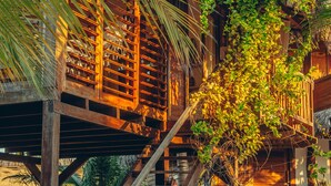 A wooden structure with a staircase leading to a thatched roof area, surrounded by lush greenery and a sandy ground. - Kitejuba Bungalows (Camocim)