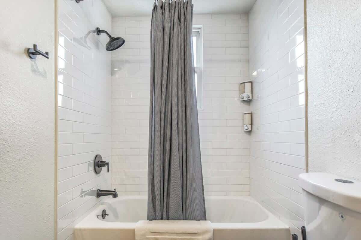 A small, modern bathroom with white tiled walls, a bathtub, a showerhead, and a curtain.