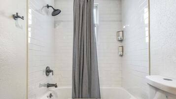 A small, modern bathroom with white tiled walls, a bathtub, a showerhead, and a curtain.