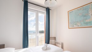 Une chambre d’hôtel avec deux lits, une grande fenêtre avec des rideaux bleus, une carte accrochée au mur et une vue sur la plage.