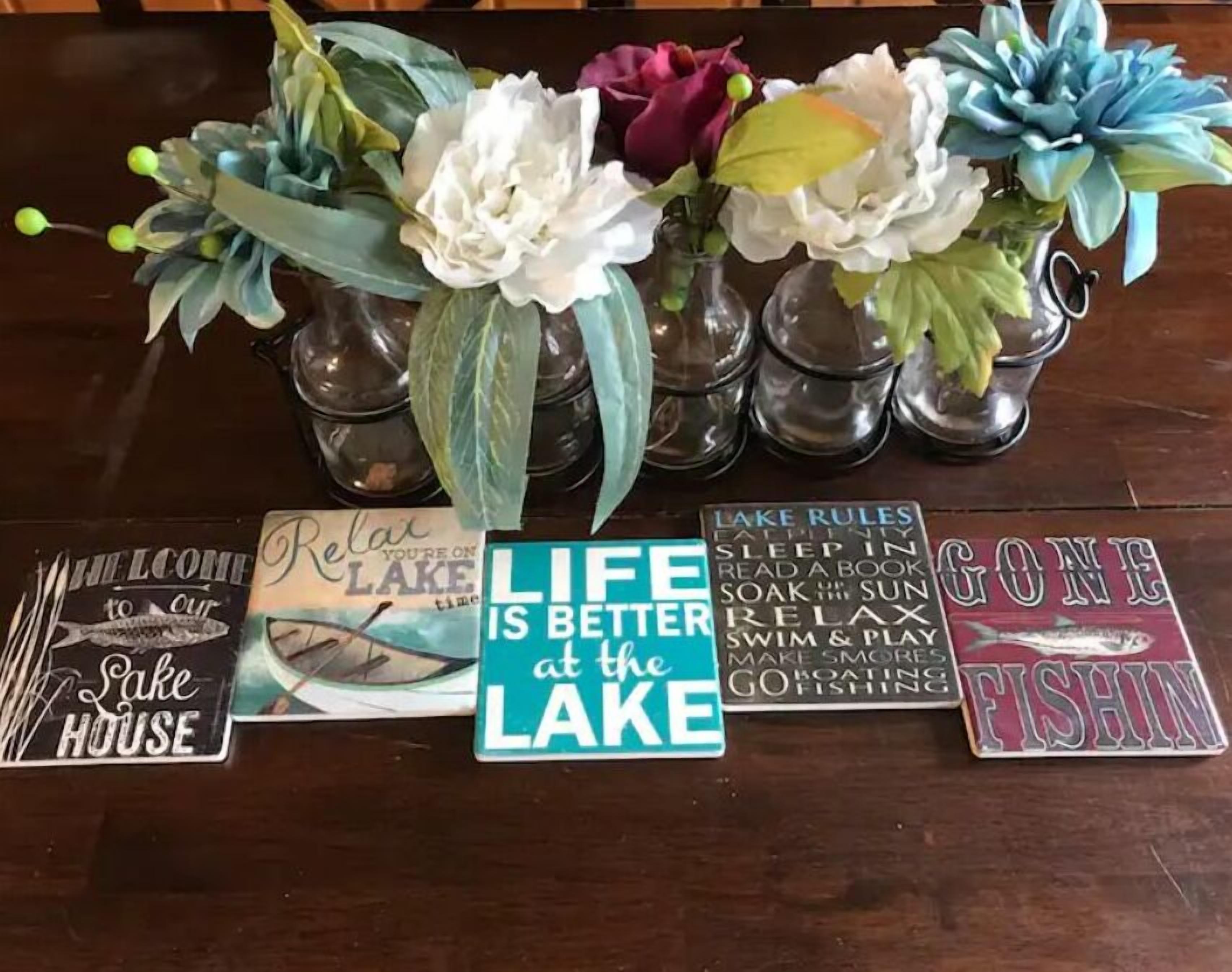 A wooden table with decorative vases containing artificial flowers and a sign that reads "LIFE IS BETTER at the LAKE".