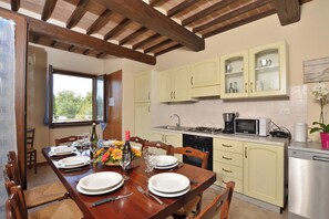 A dining area with a wooden table set for four, a kitchen with light-colored cabinets, and a window with a view of trees.