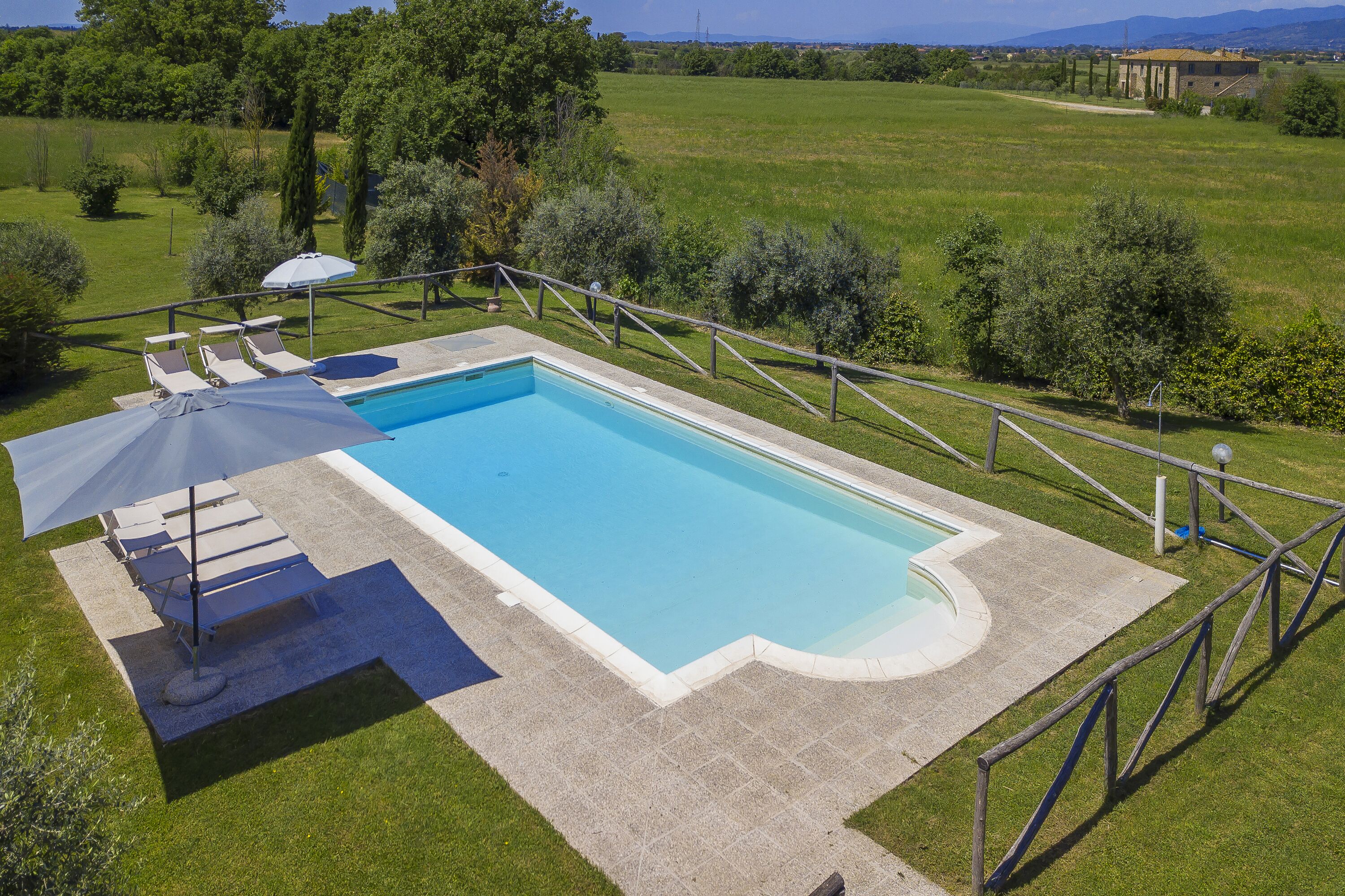 A rectangular swimming pool with a surrounding deck, a lounge area with chairs and an umbrella, and a view of a green field and distant buildings.