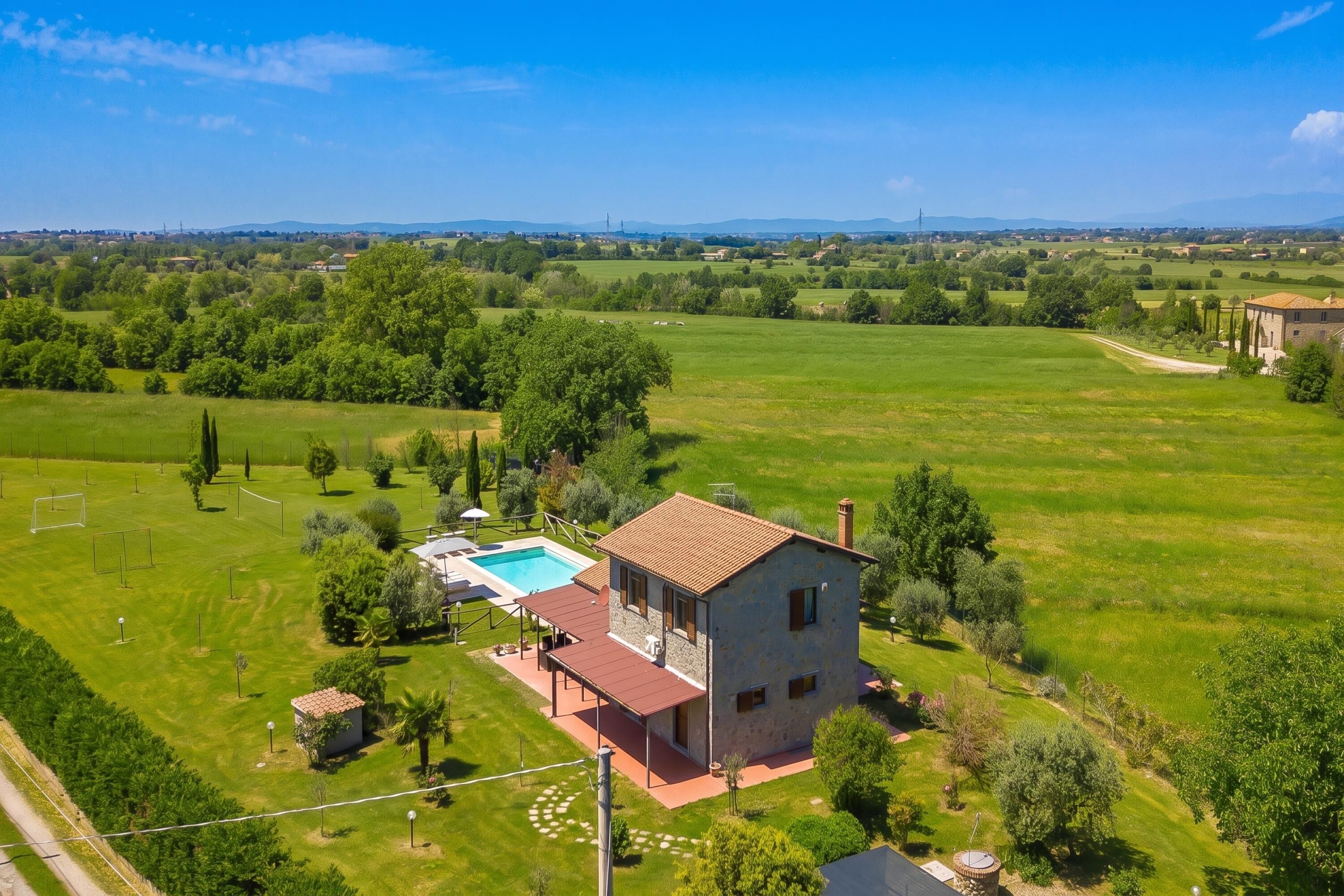 A two-story stone house with a swimming pool, surrounded by a large green field and trees.
