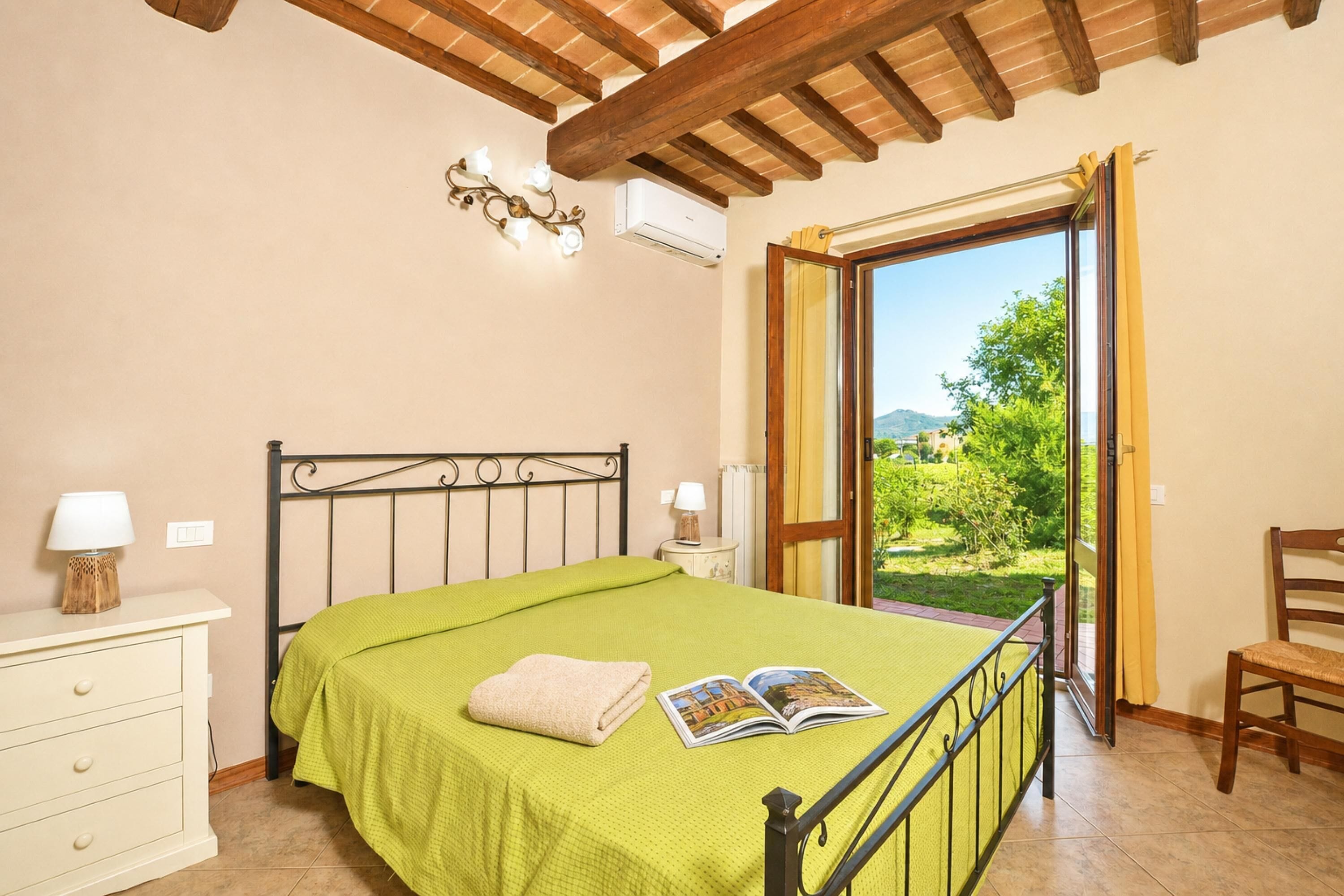 A bedroom with a bed, a nightstand, a chair, a window with a view, and a ceiling with wooden beams.