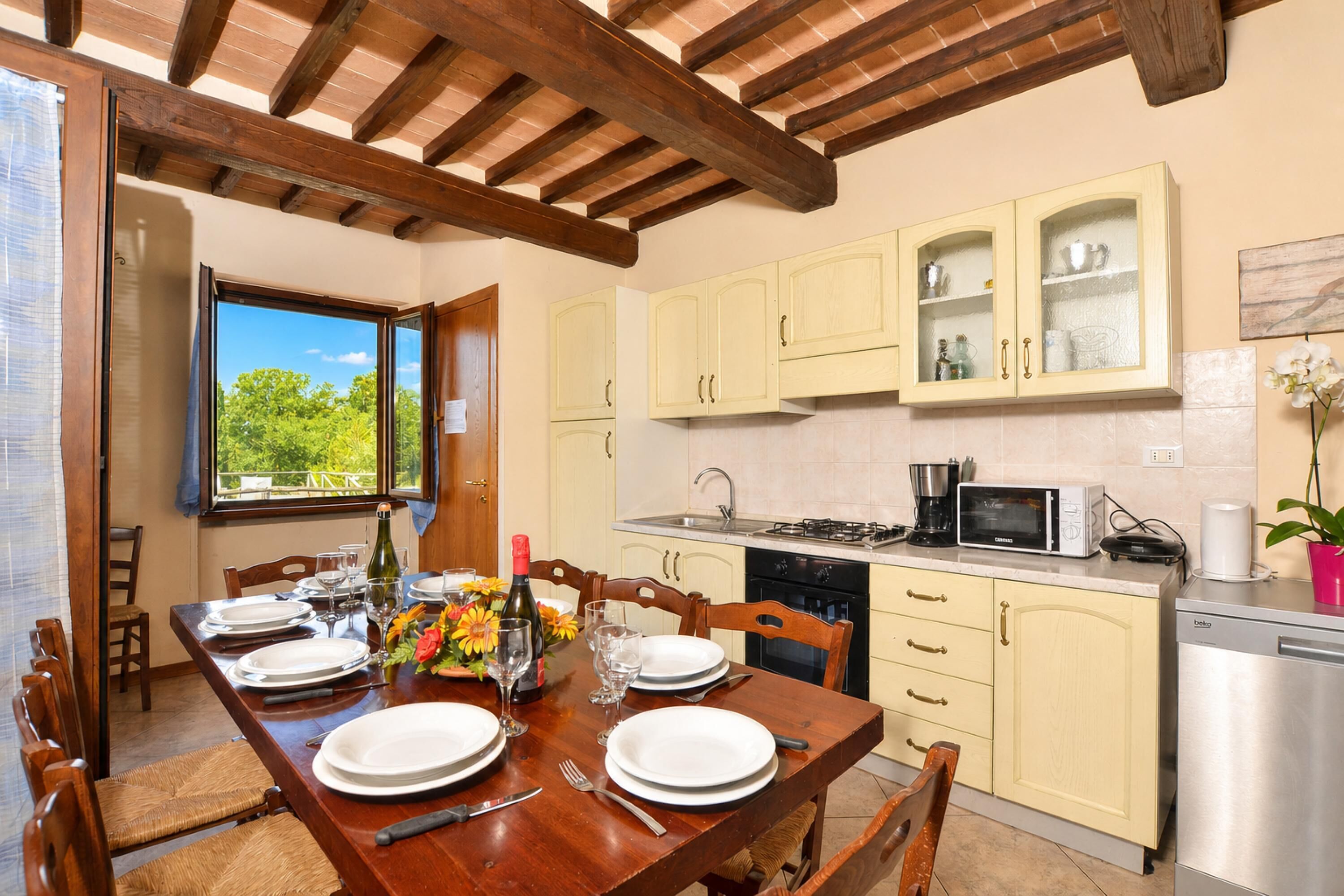 A dining area with a wooden table set for four, a kitchen with light-colored cabinets, and a view through the window.