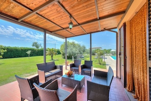 Un patio techado con mobiliario de mimbre, una mesa con un florero y vasos, y vistas a una piscina y follaje.