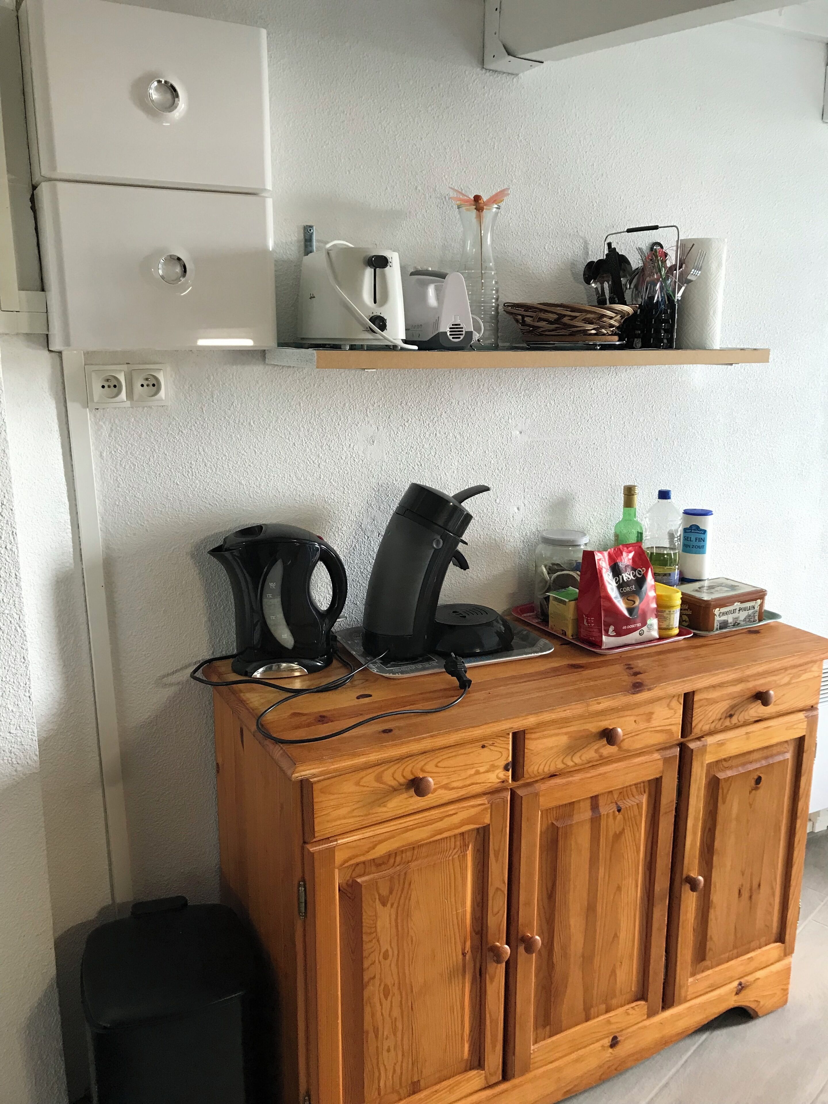 A kitchen with a wooden cabinet, a coffee maker, a kettle, and various kitchen items on the countertop.