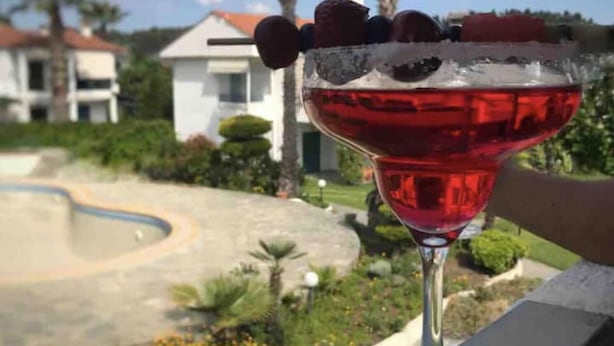 A cocktail glass with a red drink on a balcony overlooking a landscaped area with palm trees and a swimming pool. - Minimal Resort & Pool in Siviri, 150m From the Sea (Kassandra)