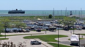 Un port de plaisance avec des bateaux, un grand navire en arrière-plan et un parking avec des voitures.