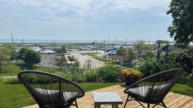 Une terrasse dotée de deux fauteuils en osier et d’une petite table, offrant une vue sur une marina avec des bateaux et une route.