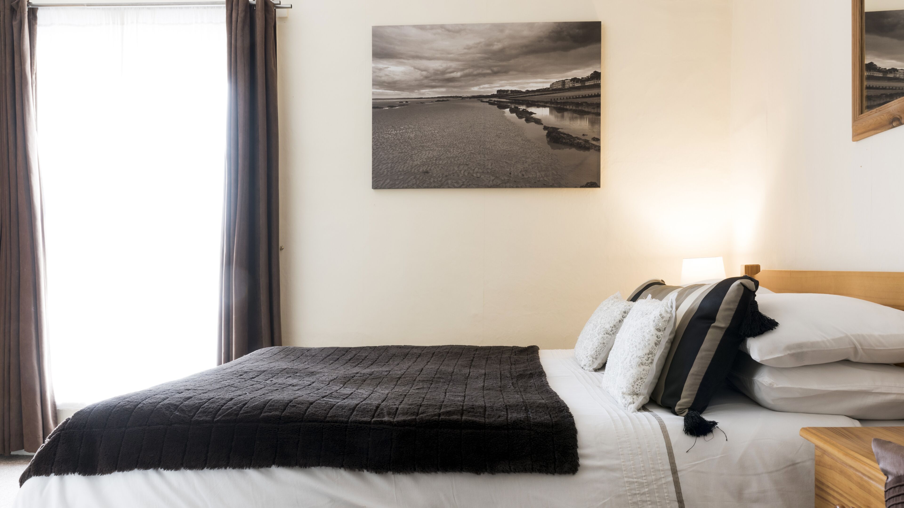 Un lit bien fait, avec une couverture et des oreillers sombres, une table de chevet avec une lampe et un tableau encadré au mur.