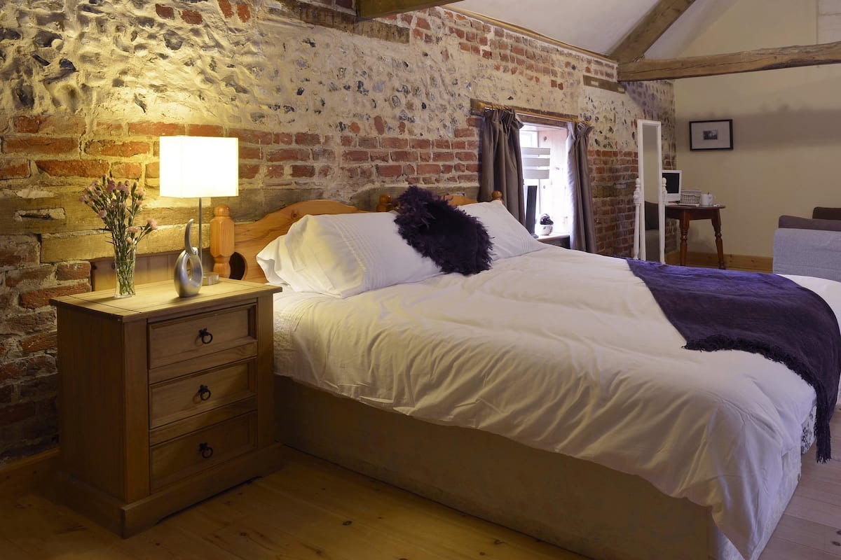 A bedroom with a wooden nightstand, a lamp, a bed with white bedding, and a brick wall.