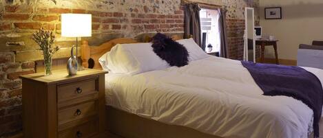 Un dormitorio con mesita de noche de madera, una lámpara, cama con ropa de cama blanca y pared de ladrillo.