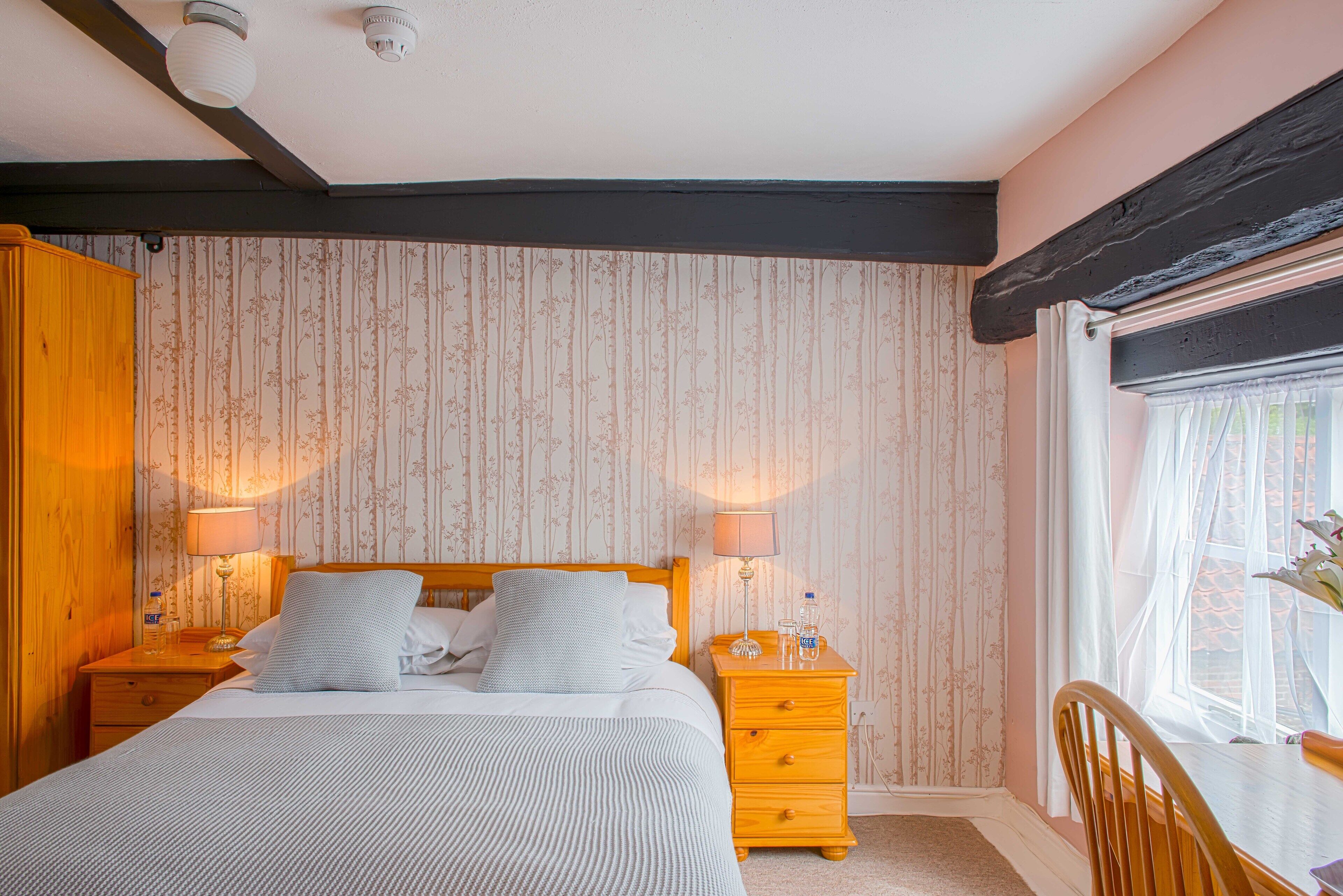 A bedroom with a bed, two bedside tables, a nightstand, a wooden wardrobe, a window with sheer curtains, and a view of greenery outside.