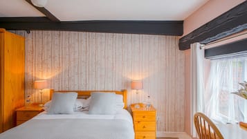 A bedroom with a bed, two bedside tables, a nightstand, a wooden wardrobe, a window with sheer curtains, and a view of greenery outside.