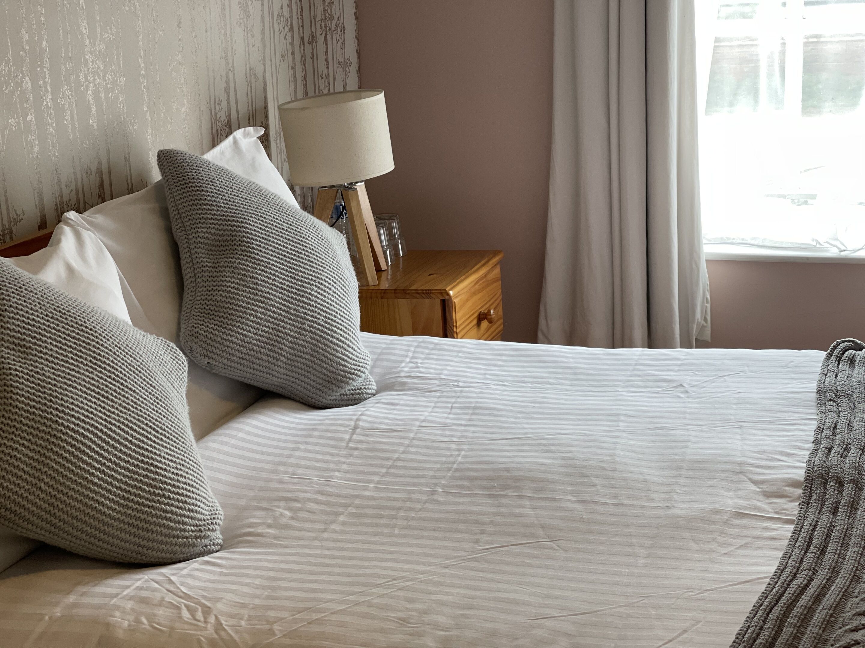 A neatly made bed with white linens and grey knitted pillows, a bedside table with a lamp, and a window with curtains.