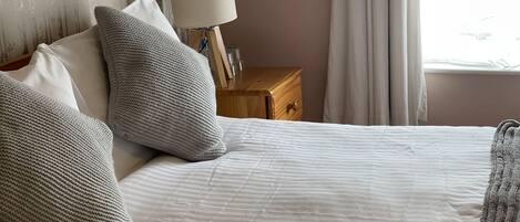 A neatly made bed with white linens and grey knitted pillows, a bedside table with a lamp, and a window with curtains.