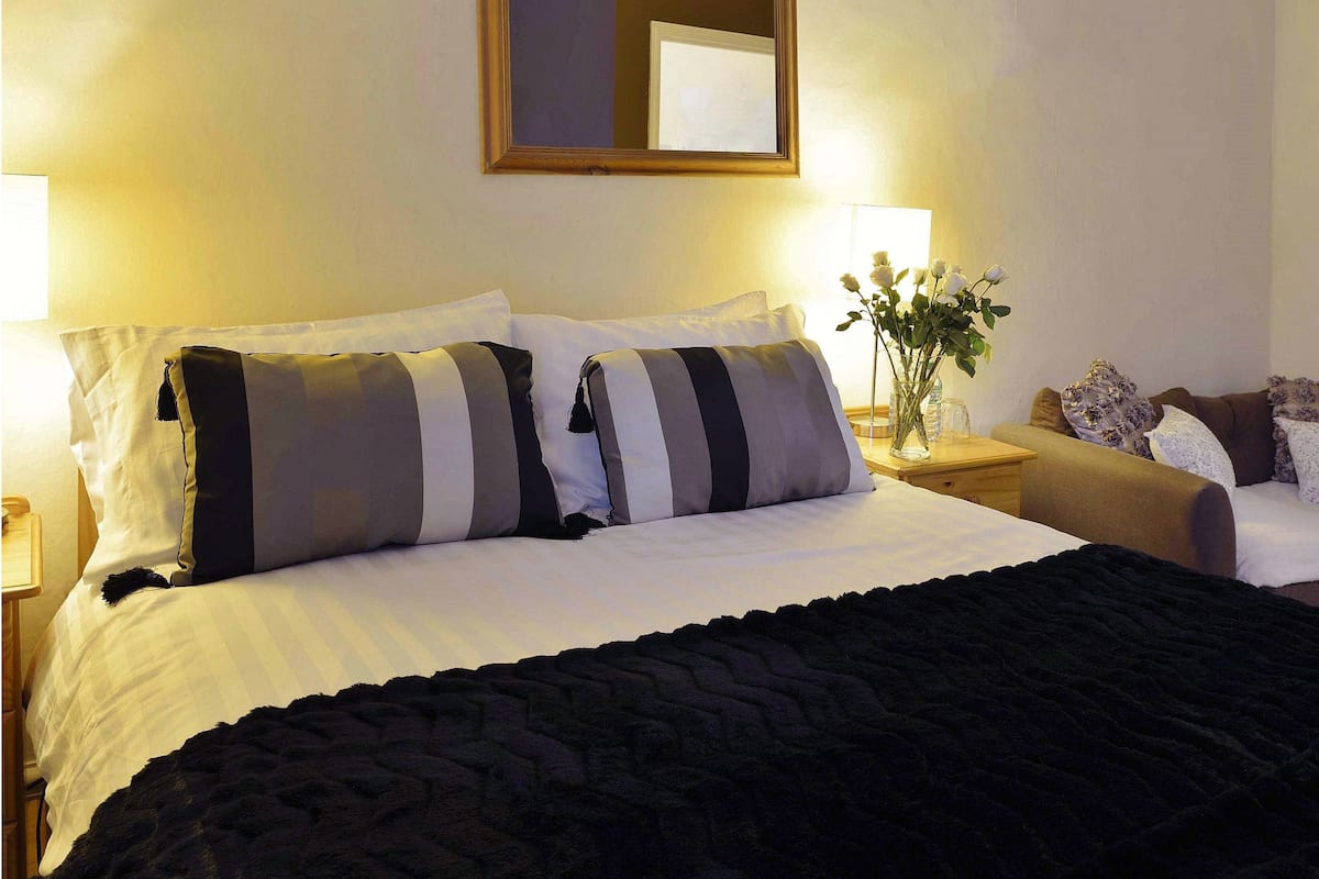 A neatly made bed with striped pillows, a dark throw blanket, and a bedside table with a vase of flowers.