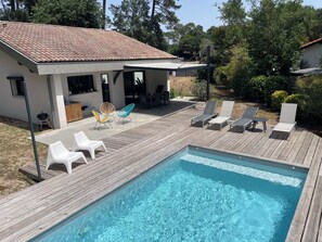 Una piscina con bordo in legno, poltrone da relax e un’area patio con barbecue.