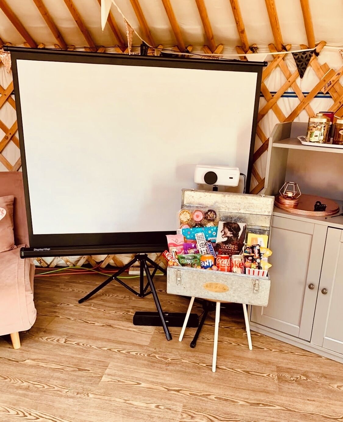 Ein gemütliches Zimmer mit einem großen Bildschirm auf einem Stativ, einem kleinen Tisch mit Snacks und einer Holzzahlende.