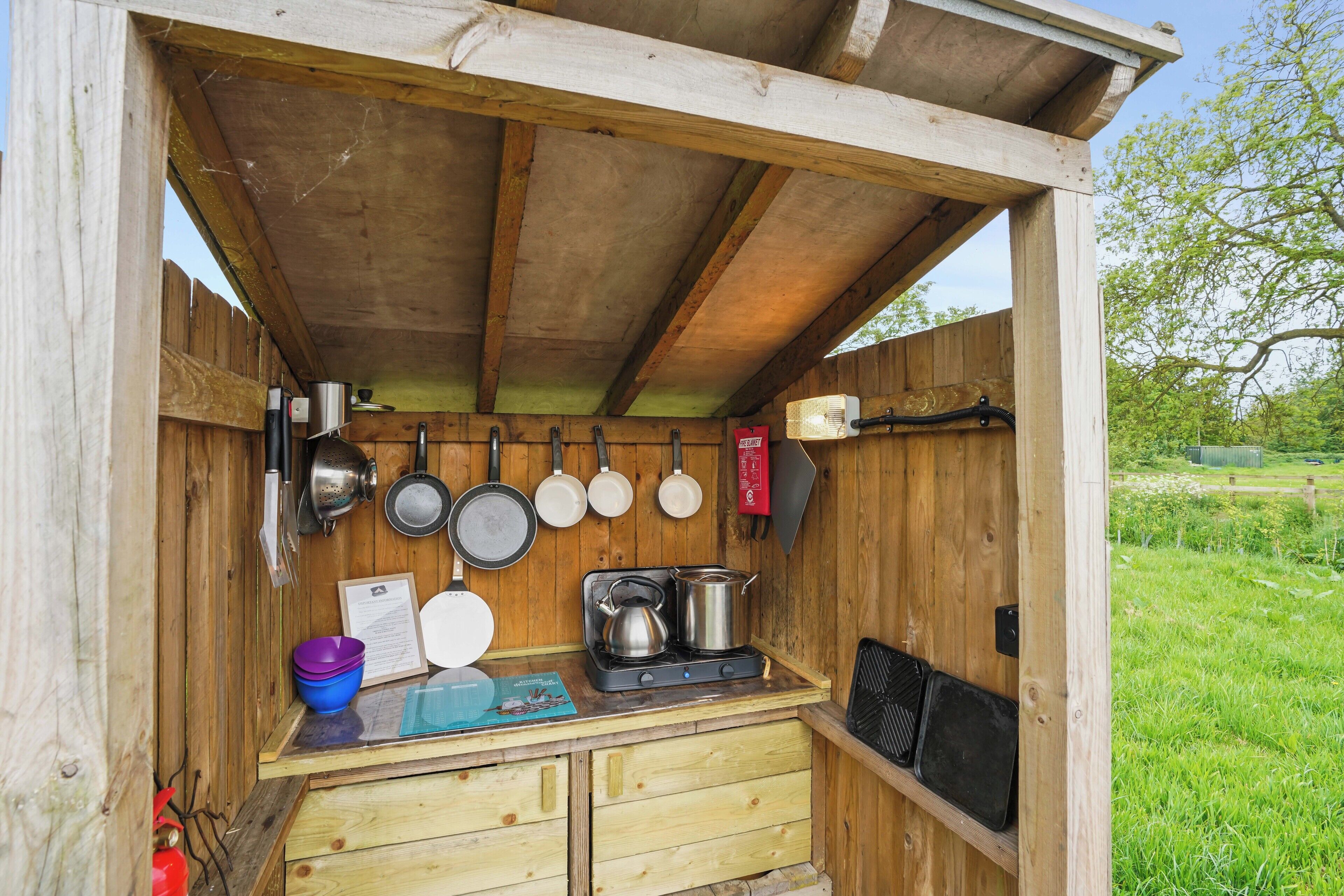 Une cuisine extérieure en bois, équipée d’une cuisinière à gaz, de casseroles, de poêles et d’une planche à découper bleue.