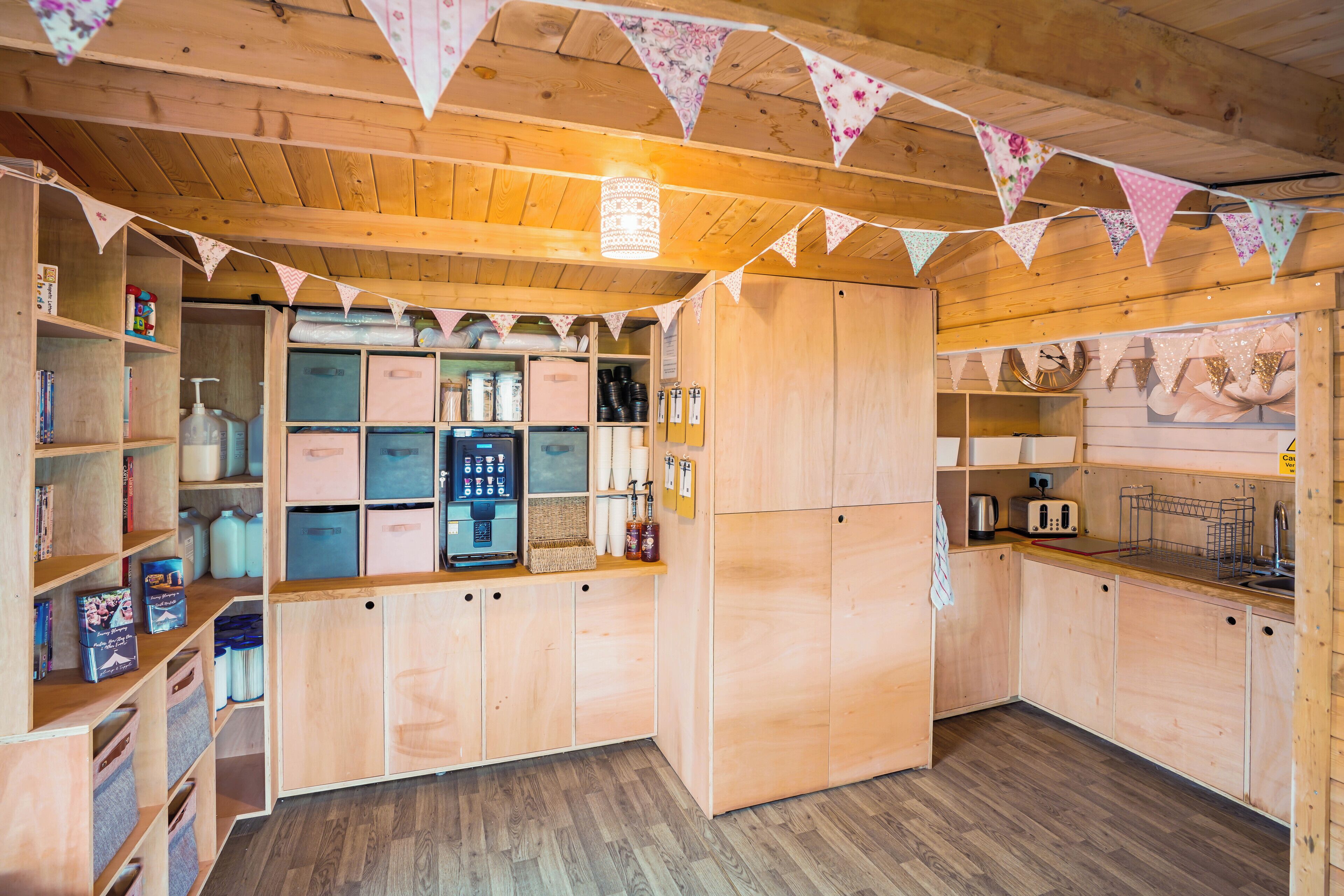 A wooden kitchen with wooden cabinets, a refrigerator, and a sink. There are shelves with various items and a window with curtains.