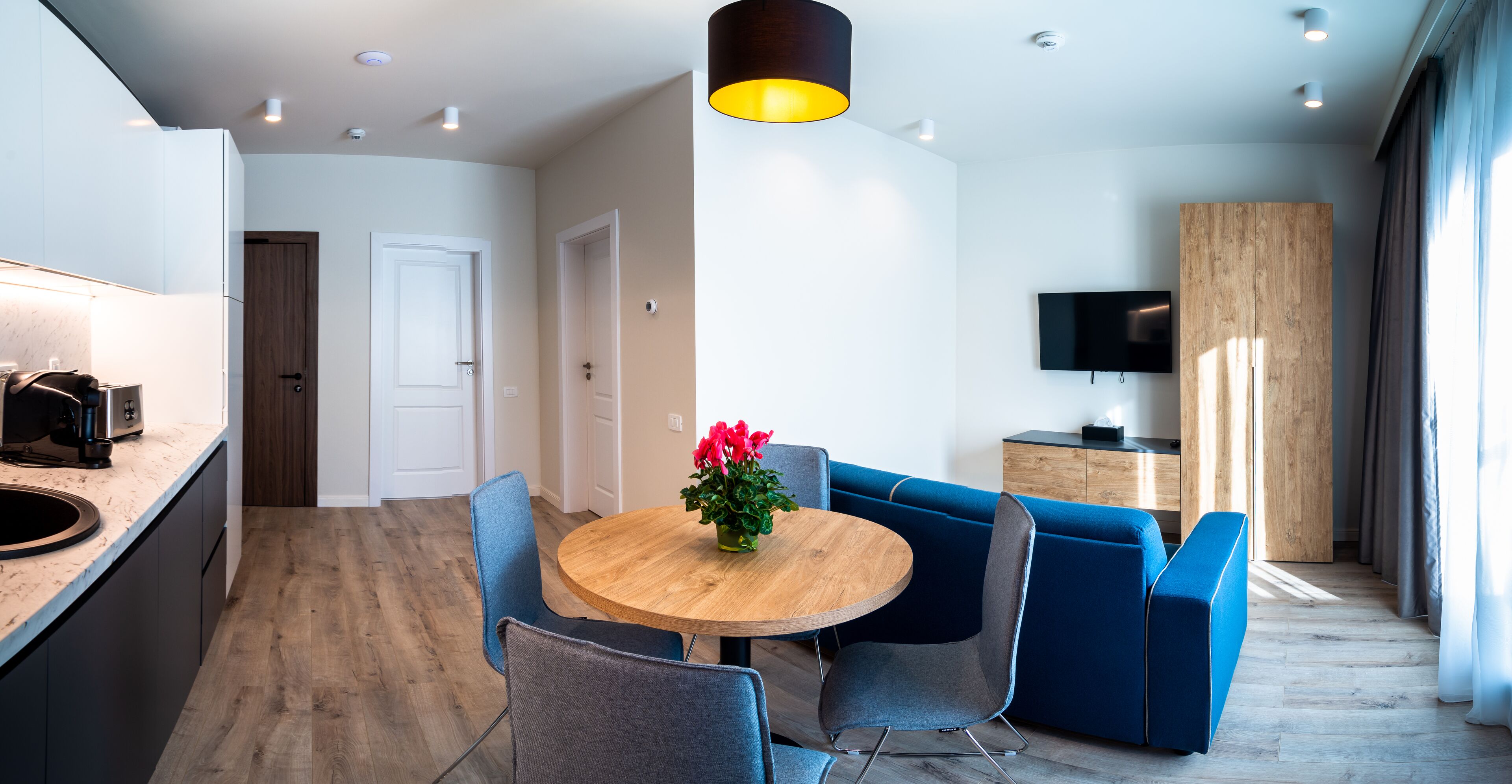 Una cocina moderna con horno empotrado y cafetera, un comedor con mesa redonda de madera y sillas azules, y una sala de estar con televisor y mueble de madera.